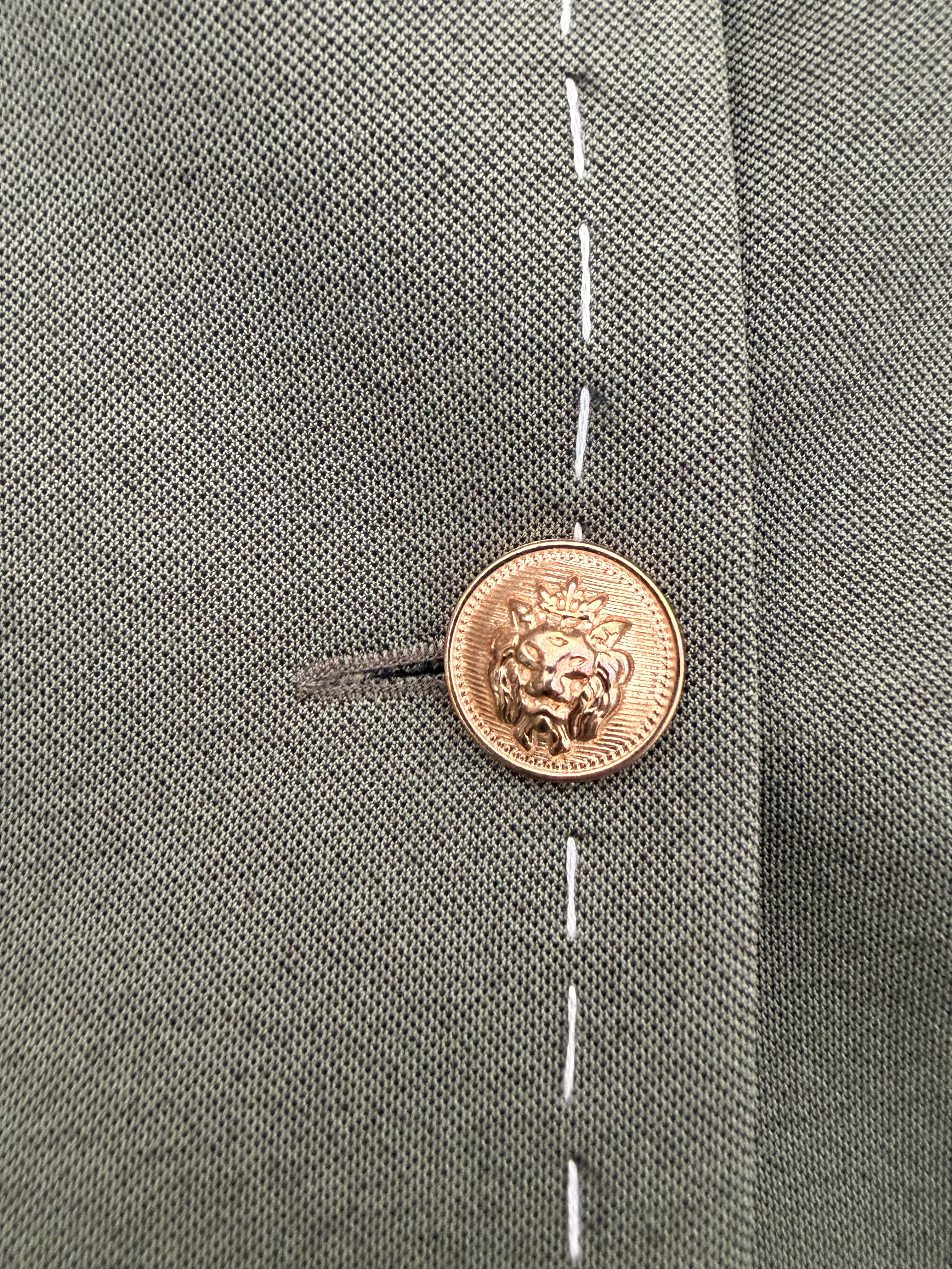 Close-up of gold lion-head button and contrast white stitching on dark olive green Debrief blazer.