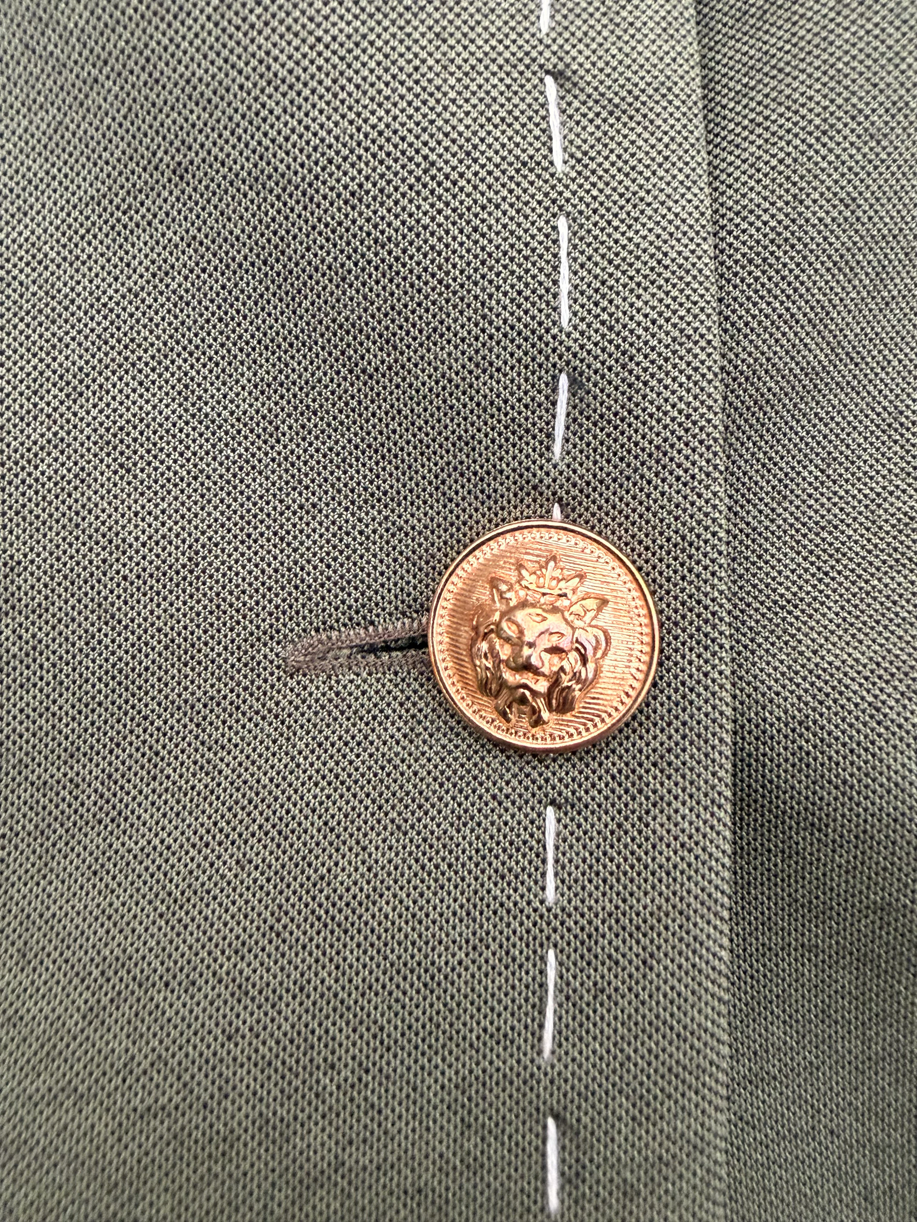 Close-up of gold lion-head button and contrast white stitching on dark olive green Debrief blazer.
